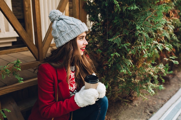 Free photo portrait pretty girl with long hair in red coat sitting on wooden stairs outdoor. she has grey knitted hat, white gloves, holds  coffee and smiling to side.