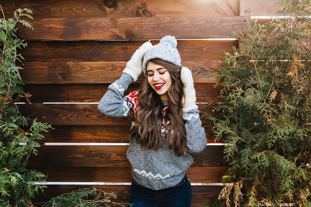 Free Photo portrait pretty girl with long hair and red lips  in knitted hat and warm gloves on wooden . she is smiling and keeps eyes closed.