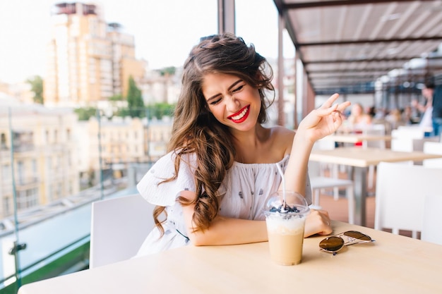 Free photo portrait of pretty girl with long hair sittting at table on the terrace in cafe. she wears a white dress with bare shoulders and red lipstick . she has a fun in front of the camera.