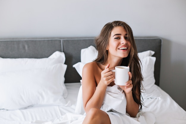 Portrait pretty girl with naughty shoulders drinking coffee on bed in the morning in apartment.