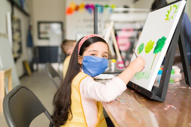 Portrait of a pretty kid wearing a face mask that is is sitting with a brush in front of a painting that she is doing in her art school