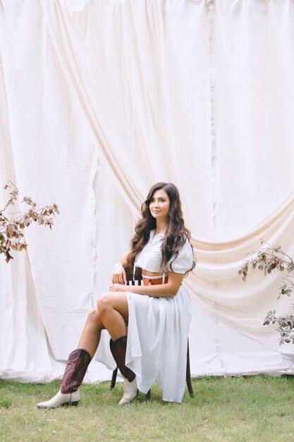 Portrait of pretty lady at garden with white wall sitting and smiling in long white dress with belt during daytime.