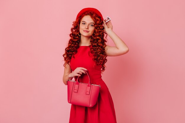 Portrait of pretty lady with wavy red hair and blue eyes. Girl in red dress puts on her beret and holds her bag.