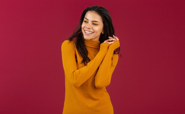 Portrait of pretty smiling stylish brunette hispanic woman in yellow autumn winter fashion dress sweater posing isolated on red wall