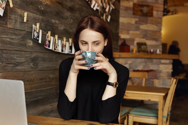 Portrait of pretty woman wearing black dress and wrist watch enjoying aroma of fresh cappuccino, holding large mug at her face while having lunch at cozy cafe surfing internet on laptop computer