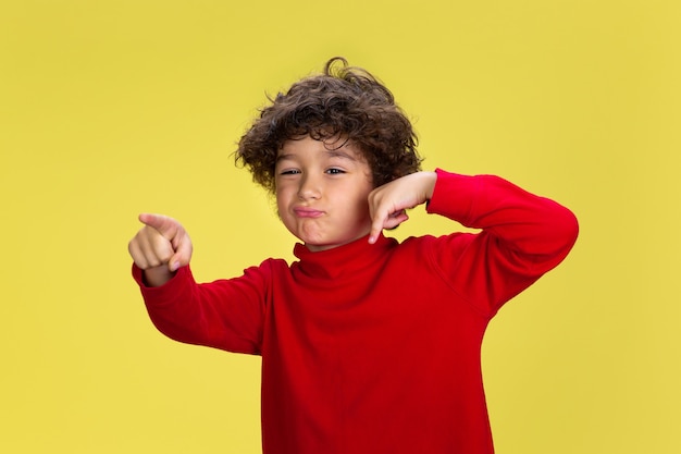 Free photo portrait of pretty young curly boy in red wear on yellow studio background. childhood, expression, education, fun concept.