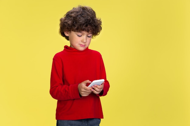 Portrait of pretty young curly boy in red wear on yellow studio background. Childhood, expression, education, fun concept.