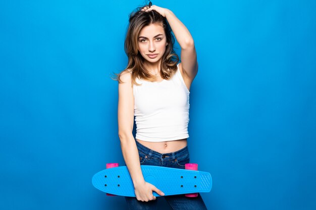 Portrait of pretty young woman in casual clothes standing,  holding skateboard isolated on blue wall . People lifestyle concept.