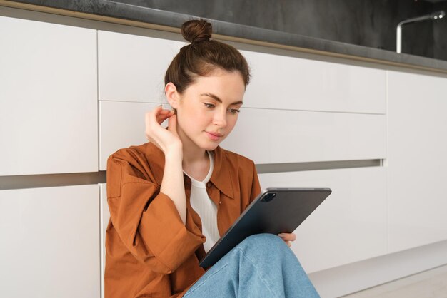 Free Photo portrait of pretty young woman at home sitting on kitchen floor looking at digital tablet using