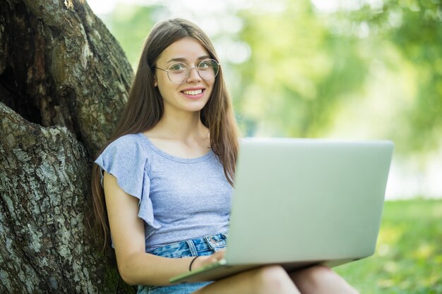 Portrait of pretty young woman sitting on green grass in park with legs crossed during summer day while using laptop