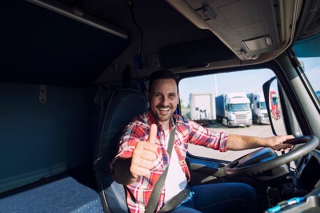 Portrait of professional motivated truck driver holding thumbs up in truck cabin