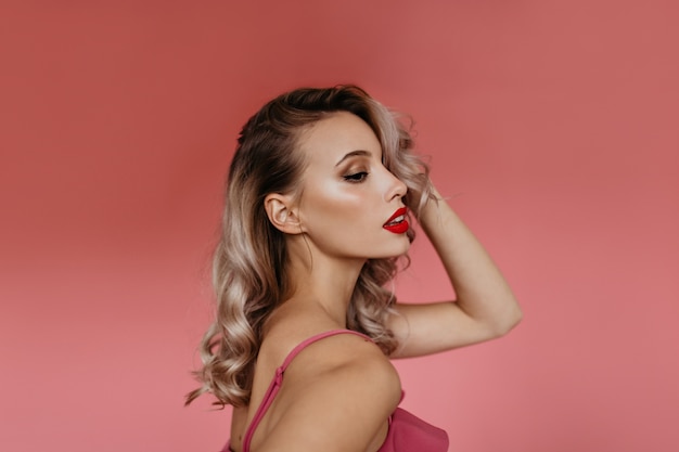 Free photo portrait in profile in studio of beautiful young blonde with curly hair and brightly painted pink lips, posing for camera showing her tender feminine shoulders