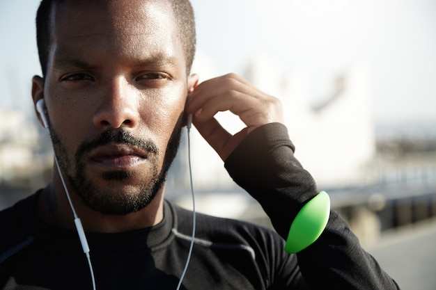 Free photo portrait of purposive fitness trainer preparing himself for tough workout. with serious face, tracker, headphones in ears african american athlete is determined to challenge himself in sport.