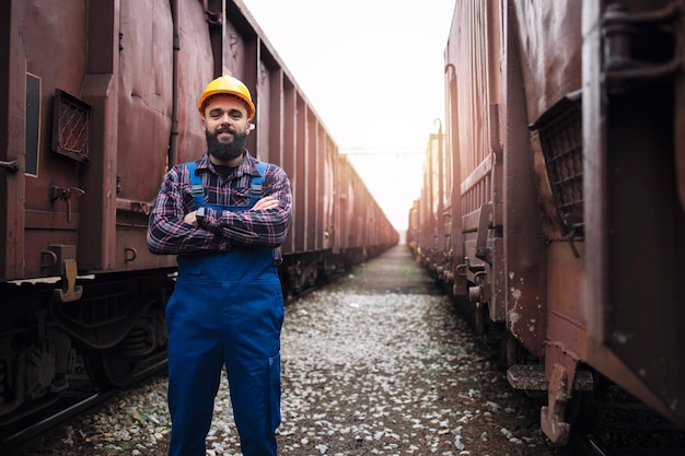 Free photo portrait of railroad worker with crossed arms proudly standing at train station between wagons