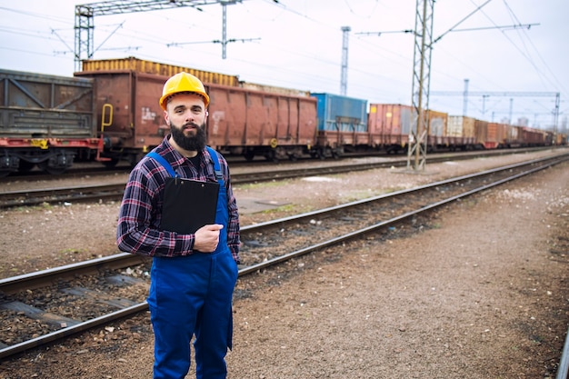 Free Photo portrait of railroader man worker with clipboard standing by the railroad tracks and cargo freight train in the background