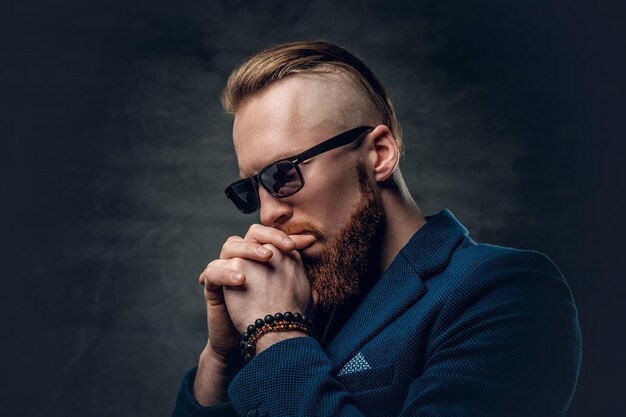 Portrait of redhead bearded hipster male dressed in a blue jacket and sunglasses over grey background.