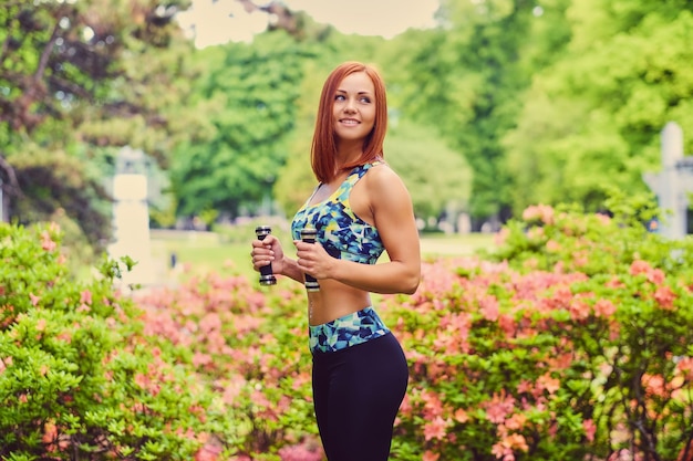 Free photo portrait of redhead fitness female holds dumbbells in a outdoor park.