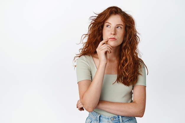 Free Photo portrait of redhead girl thinks looking aside pensive makes decision stands against white background