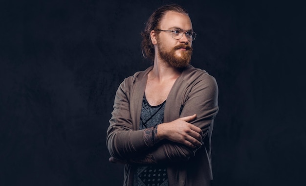 Portrait of a redhead hipster male dressed in casual clothes with glasses and full beard, standing with crossed arms in a studio. Isolated on a dark background.