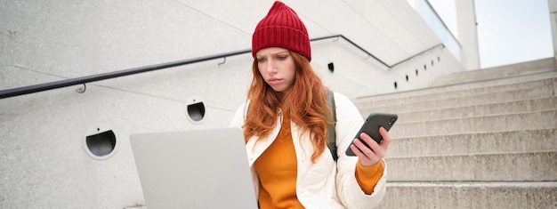 Free Photo portrait of redhead woman sits on stairs uses laptop and holds smartphone looks confused and upset