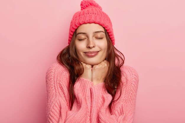 Portrait of relaxed young female model keeps both hands under chin, has eyes closed, wears rosy hat and jumper, feels satisfied, poses against pink background.