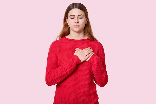 Portrait of restful impressed woman keeps both hands near heart, feels touched to hear heart piercing story, expresses good feelings