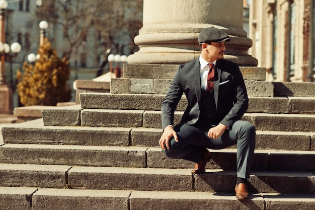 Free Photo portrait of retro 1920s english arabian business man wearing dark suit tie and flat cap near old columns