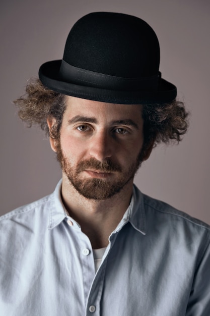 Free photo portrait of a sad looking young bearded jewish man with curly hair wearing a funny black bowler hat and light denim button up t-shirt isolated on light gray.