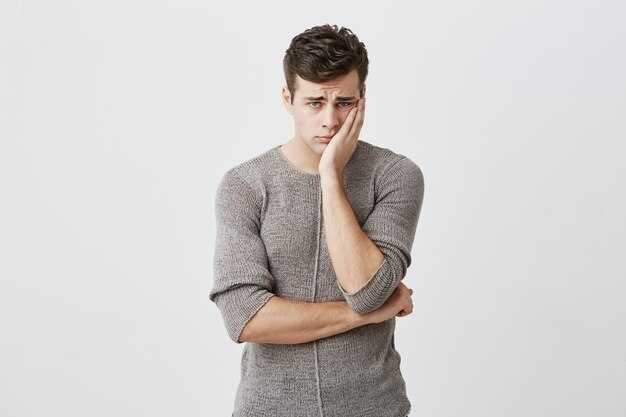 Portrait of sad upset young blue eyed male with dark hair, wearing sweater, keeping hand on cheek, frowning his face, looking with sad expression , because of bad news he received.