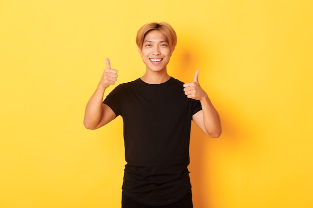 Portrait of satisfied asian man smiling and showing thumbs-up over yellow wall