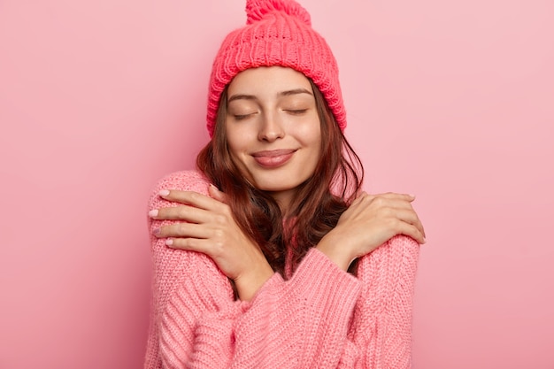 Portrait of satisfied brunette woman hugs herself, enjoys comfort in knitted warm sweater, keeps eyes closed, buys new winter outfit, isolated over pink background.