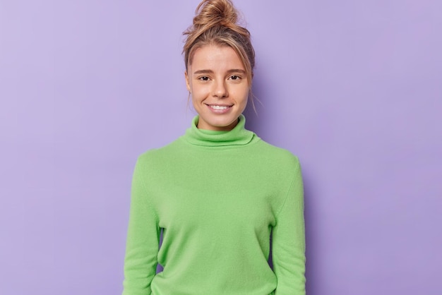 Free photo portrait of satisfied millennial girl smiles gently has healthy skin no makeup natural beauty looks directly at camera with satisfied expression wears green poloneck isolated over purple background