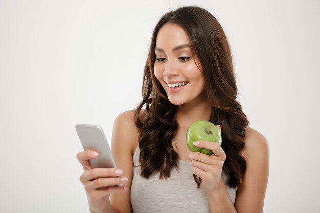 Portrait of satisfied woman using silver smartphone while eating fresh green apple, isolated over white wall