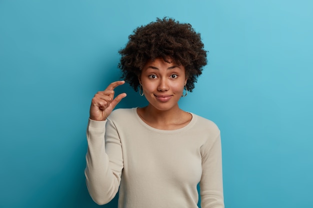 Free Photo portrait of satisfied woman with natural frizzy hair, raises hand and shapes tiny thing, tells about little size of something, says it costs little effort to gain success, wears casual white sweater