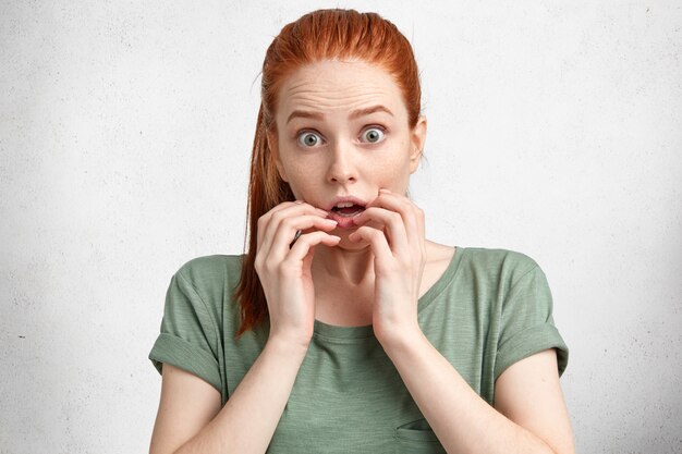 Portrait of scared beautiful red haired young woman looks with puzzlement and eyes popped out, has shocked expression, poses against white concrete