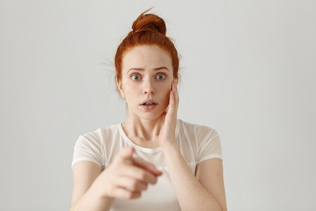 Portrait of scared bug-eyed young redhead female having fearful terrified expression, frightened with something while pointing her index finger. Danger, risk, accusation or recognition