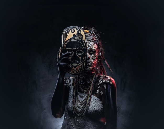 Portrait of a scary African shaman female with a petrified cracked skin and dreadlocks, holds a traditional mask on a dark background. Make-up concept.