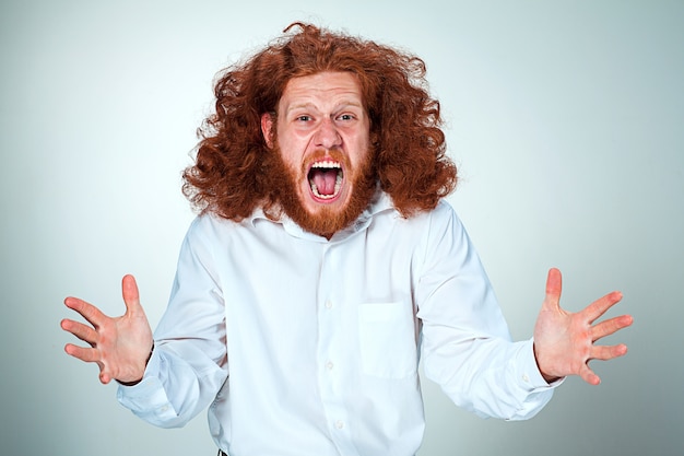 Free photo portrait of screaming young man with long red hair and shocked facial expression on gray wall