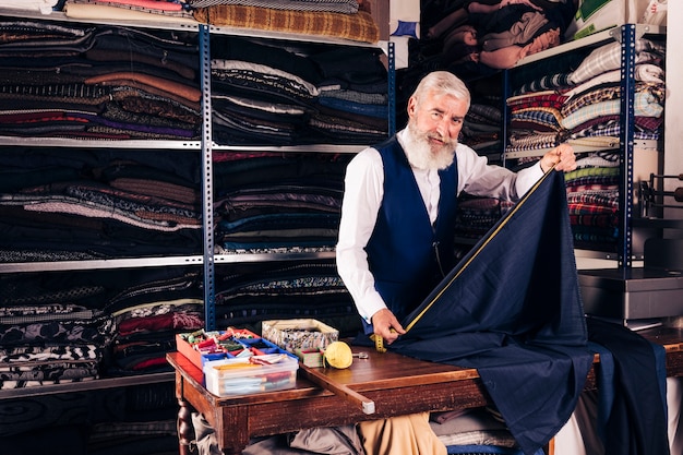 Free photo portrait of a senior male tailor taking measurement of blue fabric with measuring tape on table in his shop