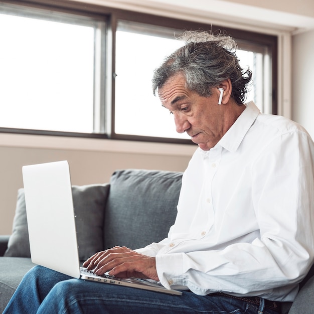 Portrait of a senior man sitting on sofa wearing bluetooth earphone using laptop