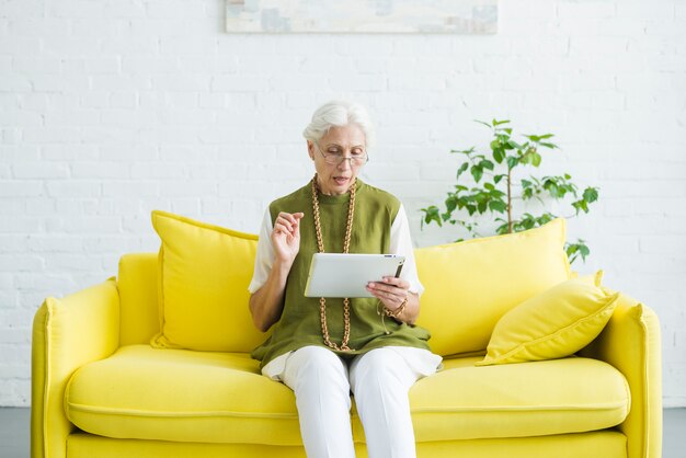 Portrait of senior woman sitting on yellow sofa looking at digital tablet