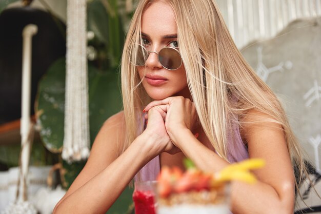 Portrait of serious blonde female with long hair, wears sunglasses, recreate at cafeteria with cocktails.