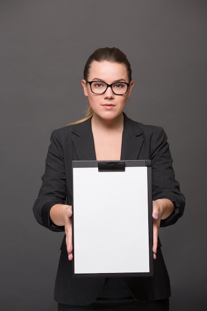 Portrait of serious businesswoman holding a clip board in front of her with blank page. Pretty lady in glasses posing in black business suit.