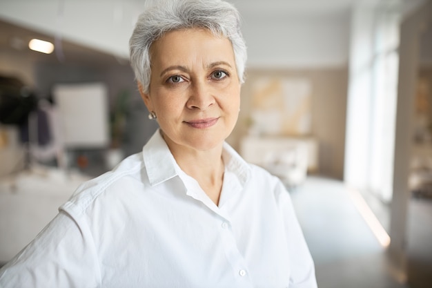 Portrait of serious confident middle aged woman with gray short hair, green eyes, wrinkles and charming smile posing indoors with arms folded