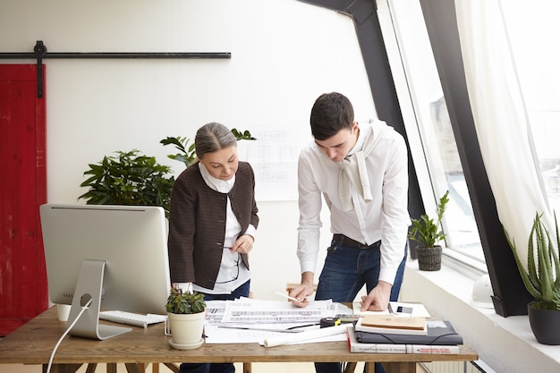 Portrait of serious creative professional designers young man and senior woman working on project, standing at office desk, creating interior designs of residential houses and commercial property