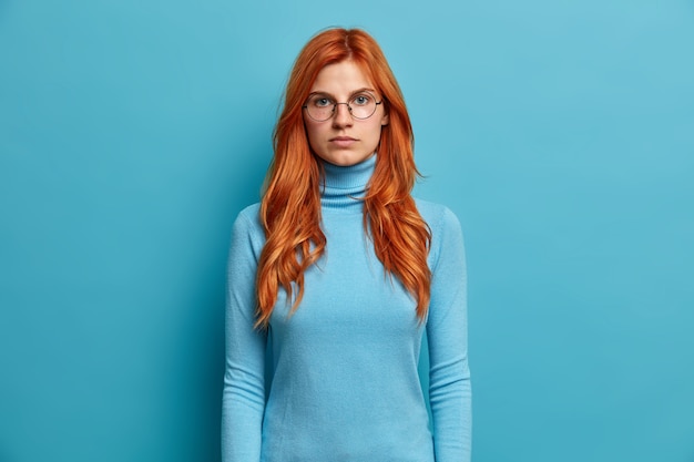 Free photo portrait of serious good looking ginger woman looks directly stands with hands down and has self confident expression dressed in blue turtleneck and spectacles.