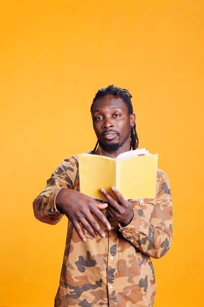 Portrait of serious reader holding literature book, reading fiction story in studio over yellow background. Concentrated african american man learning before start working at university homework