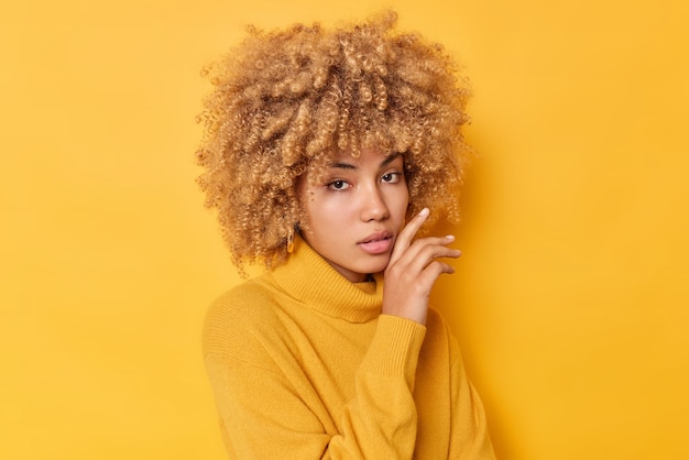 Free photo portrait of serious woman touches gently face looks directly at camer has healthy skin curly bushy hair dressed in sweater isolated over vivid yellow background human face expressions concept