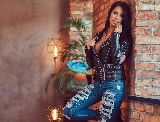 Free photo portrait of a sexy sensual brunette posing in stylish leather jacket and jeans leaning against a brick wall.