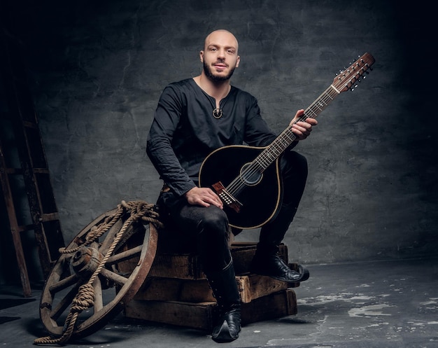 Portrait of a shaved head male musician dressed in old Celtic clothes sits on a wooden box and performing old mandolin.